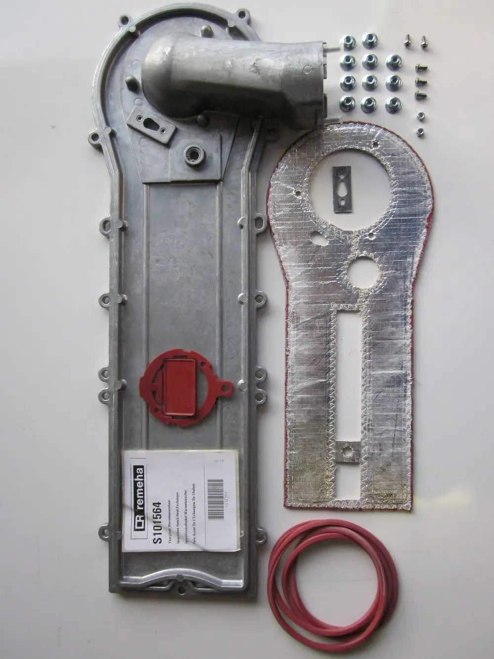 Remeha Inspektionsdeckel mit Zubehörteilen, darunter Schrauben, Dichtungen und ein Hitzeschutzblech. Teile auf einem hellen Untergrund angeordnet.